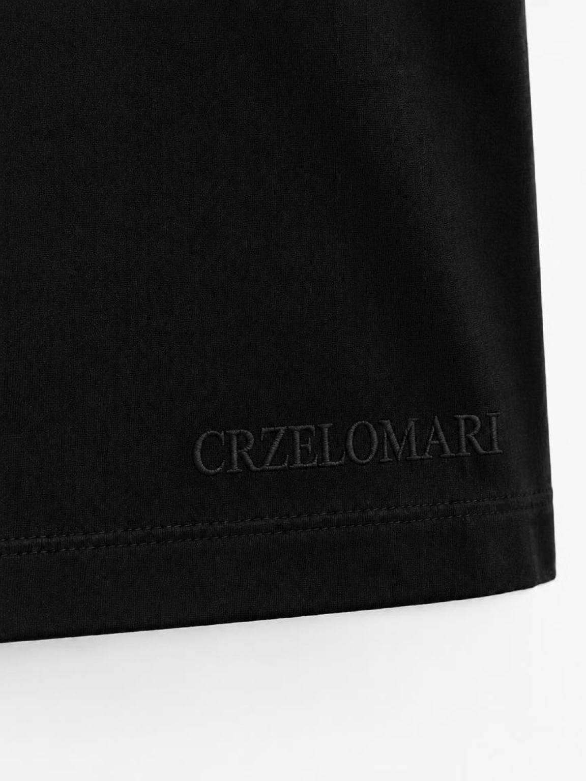 Close-up of subtle tonal CRZELOMARI embroidery on the hem of the black By Design Tee