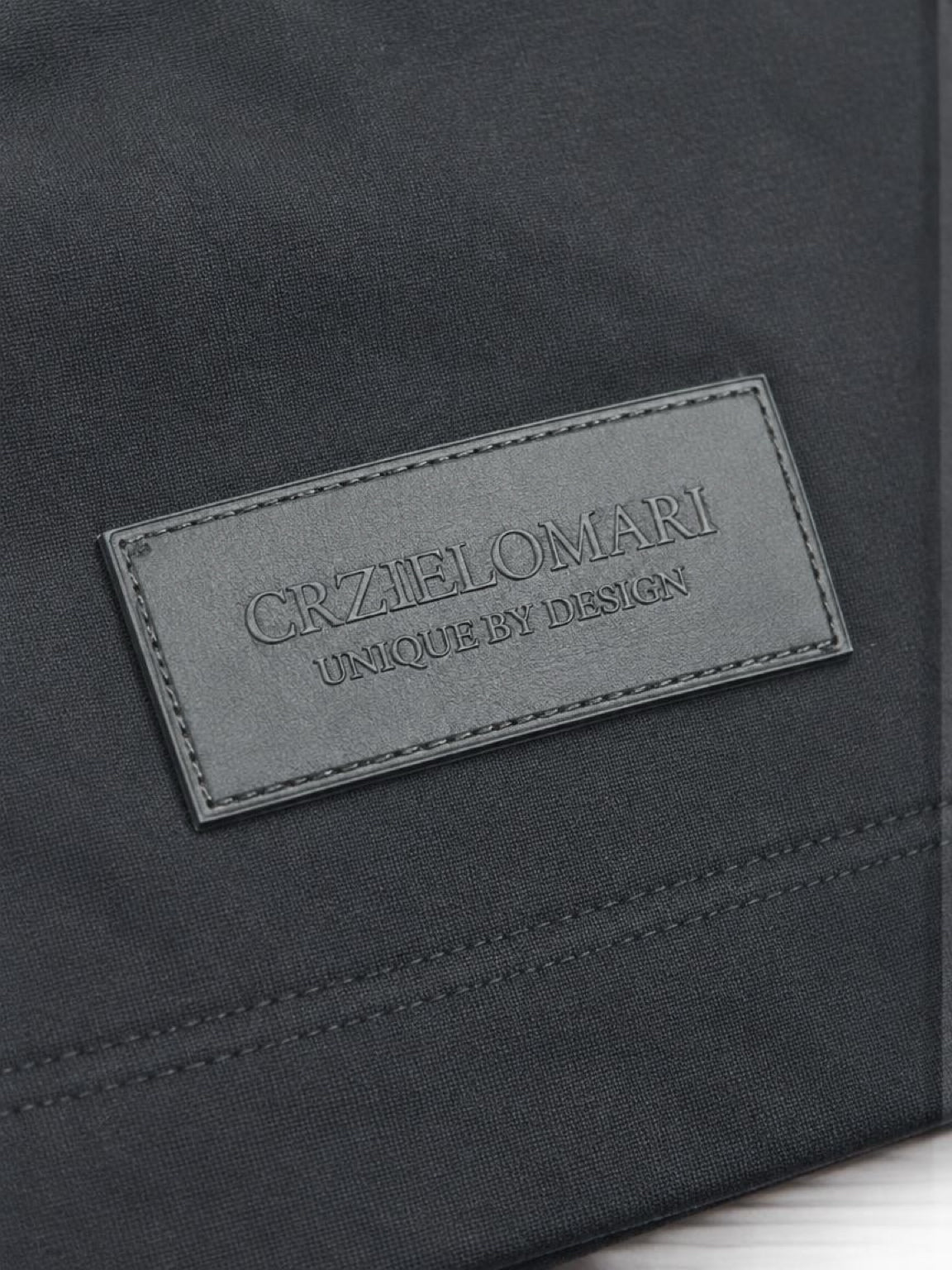 Close-up of the tonal CRZELOMARI patch on the hem of The Legacy Tee